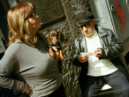 A woman and an older man pause together with a small dog on a Napoli sidewalk.